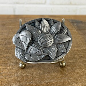 May include: A silver-coloured metal belt buckle with a detailed sunflower design and the word "KANSAS" in the centre. The buckle has a textured finish and is set against a wooden surface.