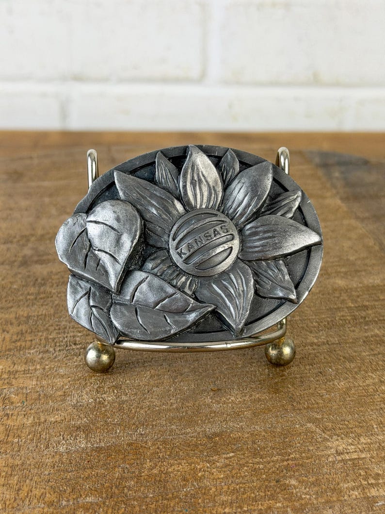 May include: A silver-coloured metal belt buckle with a detailed sunflower design and the word "KANSAS" in the centre. The buckle has a textured finish and is set against a wooden surface.