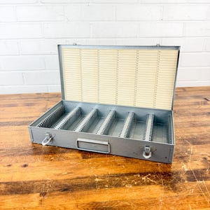 May include: A vintage gray metal storage box with multiple vertical dividers. The box has a hinged lid with a cream-colored paper insert featuring rows of small lines. A handle and latches are visible on the front.