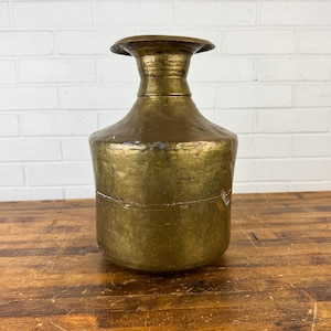 May include: A vintage brass vase with a wide base, a rounded body, and a flared top. The vase has a hammered texture and a warm, golden color. It sits on a wooden surface against a white brick wall.