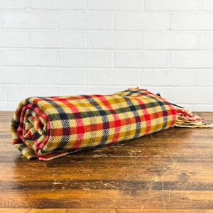 May include: A rolled-up woven blanket featuring a plaid pattern in shades of mustard yellow, bright red, and dark navy blue. The fabric has a textured appearance and includes frayed fringe edges at one end of the textile roll.
