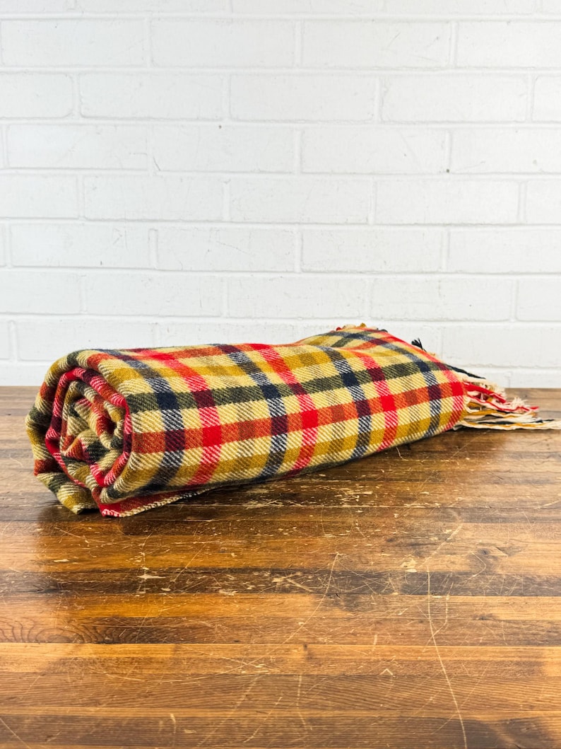 May include: A rolled-up woven blanket featuring a plaid pattern in shades of mustard yellow, bright red, and dark navy blue. The fabric has a textured appearance and includes frayed fringe edges at one end of the textile roll.