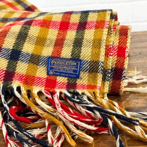 May include: A folded Pendleton wool blanket featuring a plaid pattern in mustard yellow, red, and navy blue. A blue rectangular label is stitched onto the fabric, reading Pendleton Woolen Mills, Portland, Oregon, 100% Virgin Wool. The blanket has long, multi-colored fringe.