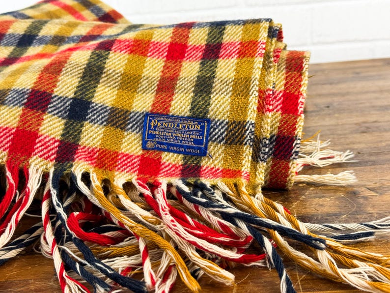 May include: A folded Pendleton wool blanket featuring a plaid pattern in mustard yellow, red, and navy blue. A blue rectangular label is stitched onto the fabric, reading Pendleton Woolen Mills, Portland, Oregon, 100% Virgin Wool. The blanket has long, multi-colored fringe.