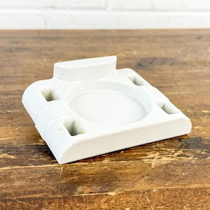 May include: A white ceramic ashtray with a rectangular base and a central circular depression. It features four small square recesses and a raised section on one side. The ashtray is placed on a wooden surface.