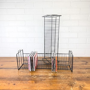 May include: Black metal CD and record storage rack. The rack has a tall, vertical section and two horizontal sections. Several CDs and records are stored in the rack. The rack is on a wooden surface in front of a white brick wall.