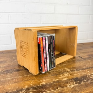 May include: A wooden crate with the text "NAPA VALLEY" and a decorative emblem. Several CDs are stacked inside the crate. The crate is made of light-colored wood and sits on a wooden surface.