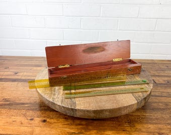 May include: A wooden box with a hinged lid, open to reveal a set of three yellow and green rulers. The box is sitting on a wooden surface.