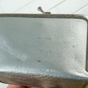 May include: A rectangular, silver-colored leather case with a metal frame and clasp. The case has a textured surface and embossed floral detailing along the bottom edge. The text "ROUND LEATHER CASE MADE IN AUSTRIA" is visible.
