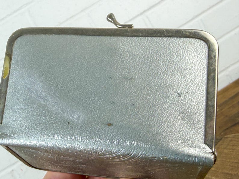 May include: A rectangular, silver-colored leather case with a metal frame and clasp. The case has a textured surface and embossed floral detailing along the bottom edge. The text "ROUND LEATHER CASE MADE IN AUSTRIA" is visible.