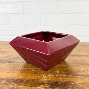 May include: A burgundy-colored, square-shaped ceramic planter with a geometric design. The planter has a glossy finish and is set on a wooden surface. The planter's sides slope inward, creating a unique, modern look. The background is a white brick wall.