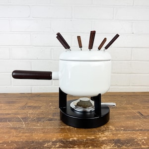 May include: A white fondue pot with a black base and a dark brown handle. The pot has a lid and several fondue forks with dark brown handles. The pot is sitting on a wooden surface.