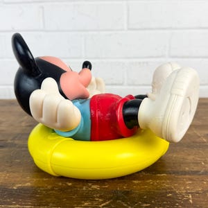 Puede incluir: Una figura vintage de Mickey Mouse en una pose relajada, con pantalones cortos rojos, una camisa azul y guantes y zapatos blancos. La figura descansa sobre un anillo inflable amarillo, con la cabeza negra y la cara rosa. La figura est&aacute; sobre una superficie de madera.