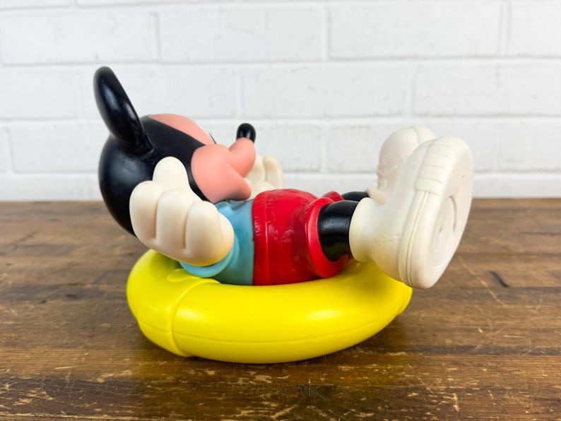 Puede incluir: Una figura vintage de Mickey Mouse en una pose relajada, con pantalones cortos rojos, una camisa azul y guantes y zapatos blancos. La figura descansa sobre un anillo inflable amarillo, con la cabeza negra y la cara rosa. La figura est&aacute; sobre una superficie de madera.