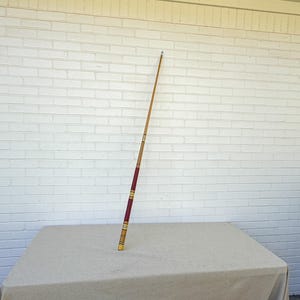 May include: A long, slender wooden pool cue standing upright. The cue features a natural light wood upper section, a dark maroon wrap area, and decorative gold-colored bands separating the different segments of the shaft and handle.