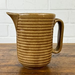 May include: A brown ceramic pitcher with a ribbed design and a handle. The pitcher is on a wooden surface.