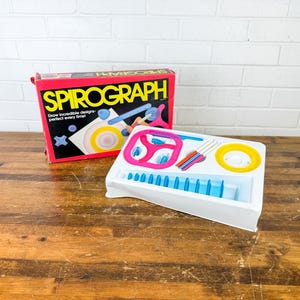 May include: A Spirograph drawing toy set with a red, black, and yellow box. The box reads "Spirograph" and "Draw incredible designs perfect every time!". The set includes a white tray with blue, pink, yellow, and orange drawing tools.
