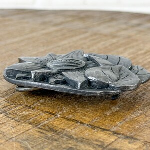 May include: A silver-coloured metal belt buckle in the shape of a sunflower. The buckle has detailed leaf and petal features, with a raised central emblem. The buckle is designed to be attached to a belt.