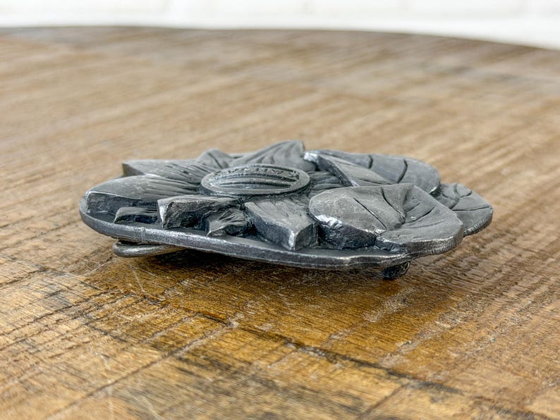 May include: A silver-coloured metal belt buckle in the shape of a sunflower. The buckle has detailed leaf and petal features, with a raised central emblem. The buckle is designed to be attached to a belt.
