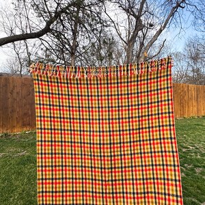 May include: A rectangular woven blanket featuring a vibrant plaid pattern of intersecting red, mustard yellow, and dark navy blue lines on a cream-colored background. The top edge of the blanket is finished with a row of short, cream-colored fringe.