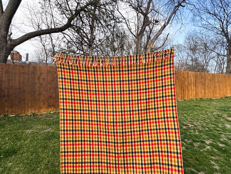 May include: A rectangular woven blanket featuring a vibrant plaid pattern of intersecting red, mustard yellow, and dark navy blue lines on a cream-colored background. The top edge of the blanket is finished with a row of short, cream-colored fringe.