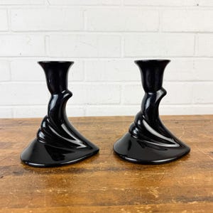 May include: Two black, glossy candle holders with a unique, twisted design. The candle holders have a flared base and a cup-shaped top. They are placed on a wooden surface against a white brick wall.