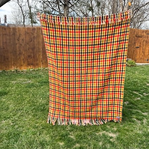 May include: A rectangular woven blanket featuring a vibrant plaid pattern in shades of mustard yellow, deep red, and dark navy blue. The textile has a consistent grid design throughout and is finished with decorative fringe along the top and bottom edges.
