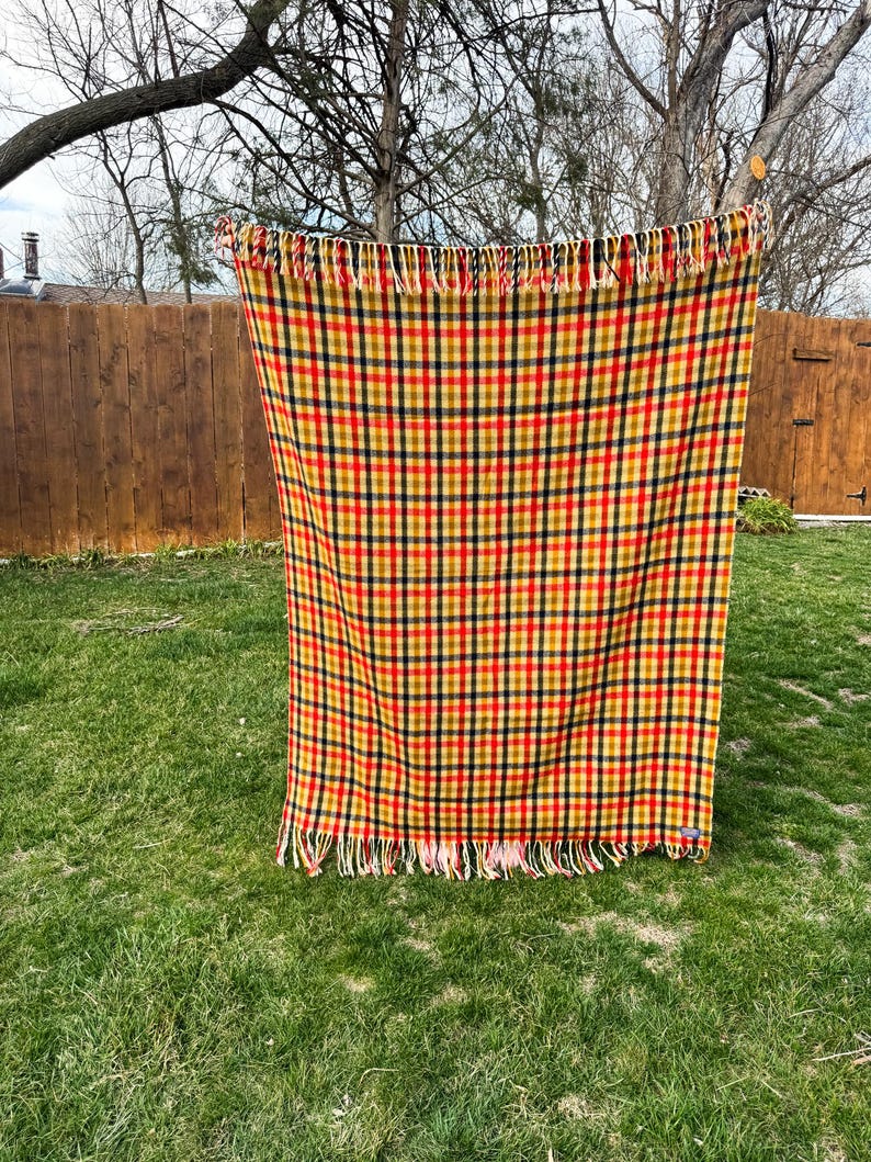 May include: A rectangular woven blanket featuring a vibrant plaid pattern in shades of mustard yellow, deep red, and dark navy blue. The textile has a consistent grid design throughout and is finished with decorative fringe along the top and bottom edges.
