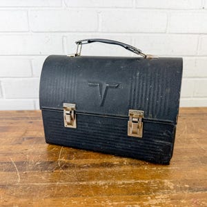 May include: A vintage black lunchbox with a handle and two metal clasps. The lunchbox has a textured surface with vertical lines and a stylized "T" emblem on the front. The lunchbox is sitting on a wooden surface.