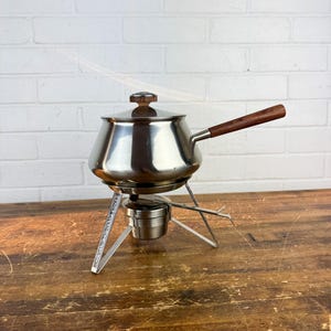 May include: A vintage stainless steel fondue pot with a wooden handle and lid knob. The pot sits on a metal stand with a fuel burner below. The background is a wooden surface and a white brick wall.