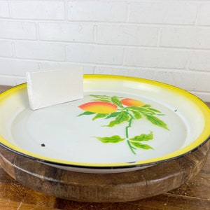 Puede incluir: Una bandeja de servir de esmalte blanco con un borde amarillo y un dise&ntilde;o pintado de hojas verdes y frutas naranjas. La bandeja est&aacute; sentada sobre una base de madera.