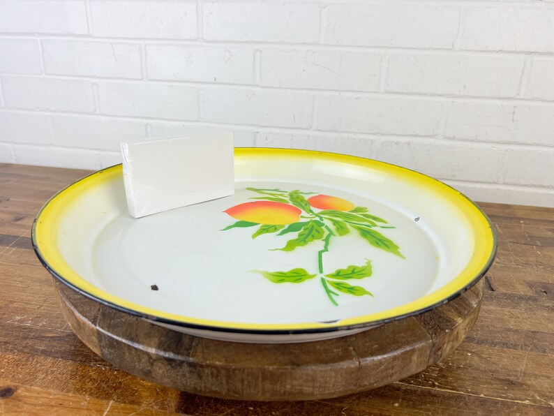 Puede incluir: Una bandeja de servir de esmalte blanco con un borde amarillo y un dise&ntilde;o pintado de hojas verdes y frutas naranjas. La bandeja est&aacute; sentada sobre una base de madera.