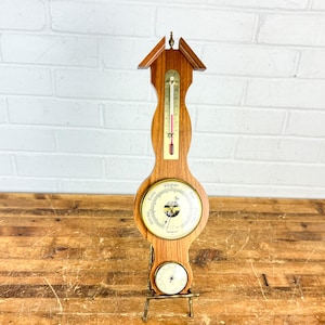 May include: A wooden weather station with a thermometer, barometer, and hygrometer. The thermometer has a red liquid column. The barometer has a white face with black markings. The hygrometer is at the bottom. The weather station is mounted on a brass stand.