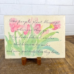May include: A white ceramic tile with a watercolor painting of pink flowers and green leaves. The tile has a quote that reads: "Some people plant blessings wherever they go - They leave behind smiles that blossom for miles and more gratitude than they know."