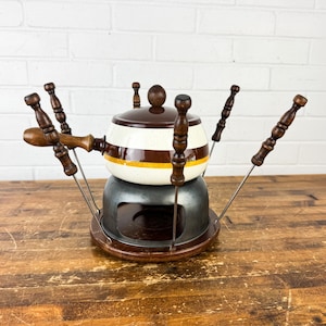 May include: A vintage fondue set with a brown and white ceramic pot, a metal stand, and wooden-handled forks. The pot has a brown lid and a yellow stripe. The set is on a wooden surface.