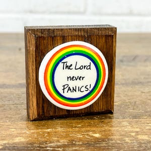 May include: A small, square wooden block with a round button affixed to the front. The button has a white background with a rainbow border and the text "The Lord never PANICS!" in black.