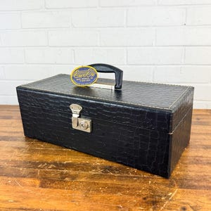 May include: Black rectangular case with a textured, crocodile-like pattern. It features a silver clasp, a black handle, and a tag that reads "Ess & Ess Music Products Inc." The case is on a wooden surface.