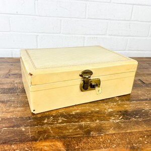May include: A rectangular, cream-colored box with a brass clasp and keyhole. The box has a textured lid and sits on a wooden surface. The box is closed and appears to be vintage.