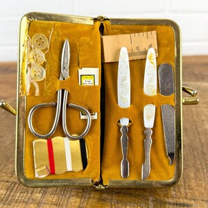 May include: Open vintage sewing kit with a gold-tone frame and mustard-yellow interior. The kit contains scissors, buttons, needles, a ruler, and various manicure tools. The tools have decorative floral designs.