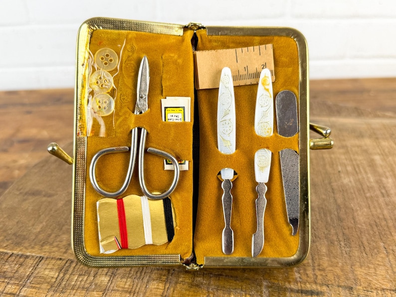 May include: Open vintage sewing kit with a gold-tone frame and mustard-yellow interior. The kit contains scissors, buttons, needles, a ruler, and various manicure tools. The tools have decorative floral designs.