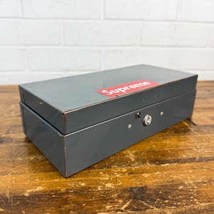 May include: A gray metal box with a red and white "Supreme" sticker on the lid. The box has a lock and keyhole on the front. The box is sitting on a wooden surface.