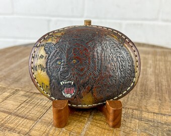 May include: A brown leather belt buckle featuring a detailed carving of a bear with a snarling expression. The bear is depicted in shades of brown, black, and yellow.