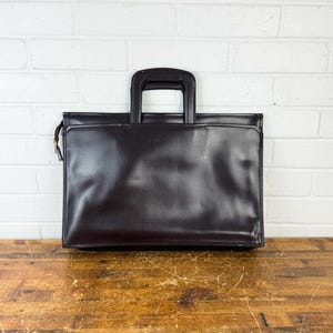 May include: A dark brown leather briefcase with a top handle and a zipper closure. The briefcase is rectangular and sits on a wooden surface. The background is a white brick wall.