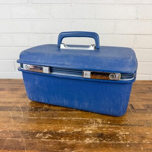 May include: A vintage blue travel case with a textured surface, a silver-toned clasp, and a matching blue handle. The case is rectangular and sits on a wooden surface.