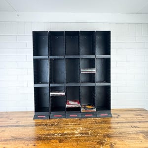 May include: Black CD storage rack with multiple compartments. The rack is made of black plastic and has a rectangular shape. Some compartments contain CDs. The rack is on a wooden table against a white brick wall.