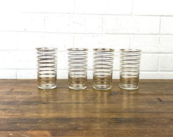 May include: Four clear drinking glasses with gold horizontal stripes. The glasses have a cylindrical shape and are arranged in a row on a wooden surface. The background is a white brick wall.