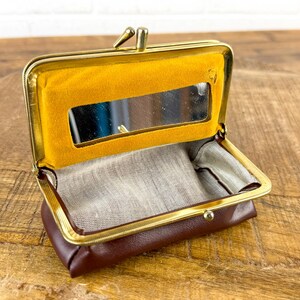 May include: Open brown leather coin purse with a gold-colored frame. The interior features a small mirror and a yellow felt lining. The purse is open, revealing a light-colored fabric lining.