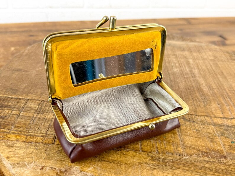May include: Open brown leather coin purse with a gold-colored frame. The interior features a small mirror and a yellow felt lining. The purse is open, revealing a light-colored fabric lining.
