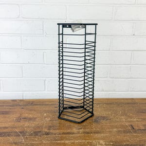 May include: A tall, black wire storage rack with a hexagonal base and vertical sides. The structure consists of thin metal bars arranged in a grid pattern, featuring a small mounting bracket at the top center for wall attachment purposes.