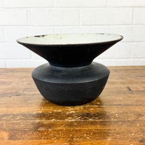 May include: A black ceramic vase with a wide, flared top and a rounded base. The interior of the top is white, contrasting with the dark exterior. The vase sits on a wooden surface.